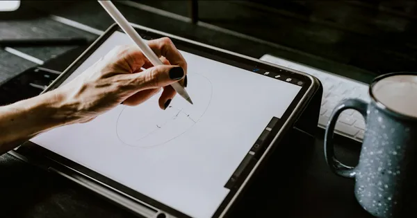 Main tenant un stylet et dessinant sur une tablette, à côté d'un clavier et d'une tasse